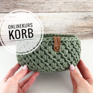 以下が含まれることがあります： 茶色の革の取っ手が付いた緑色の編み物のバスケット。バスケットは2つの手で持たれています。背景には「ONLINEKURS KORB」というテキストが表示されています。