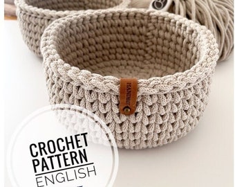 May include: Two beige crocheted baskets with a brown leather tag that says "Handmade". The baskets are on a wooden surface. Crochet pattern in English.