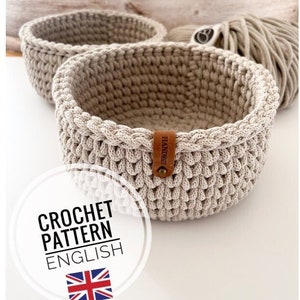 Pode incluir: Duas cestas de crochê bege com uma etiqueta de couro marrom que diz "Handmade". As cestas estão sobre uma superfície de madeira. Padrão de crochê em inglês.