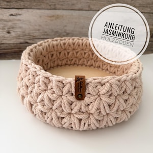 May include: A round, light beige crocheted basket featuring a star-like stitch pattern and a smooth wooden base. A small brown leather tag is attached to the side. The text ANLEITUNG JASMINKORB HOLZBODEN is visible in a circular overlay.