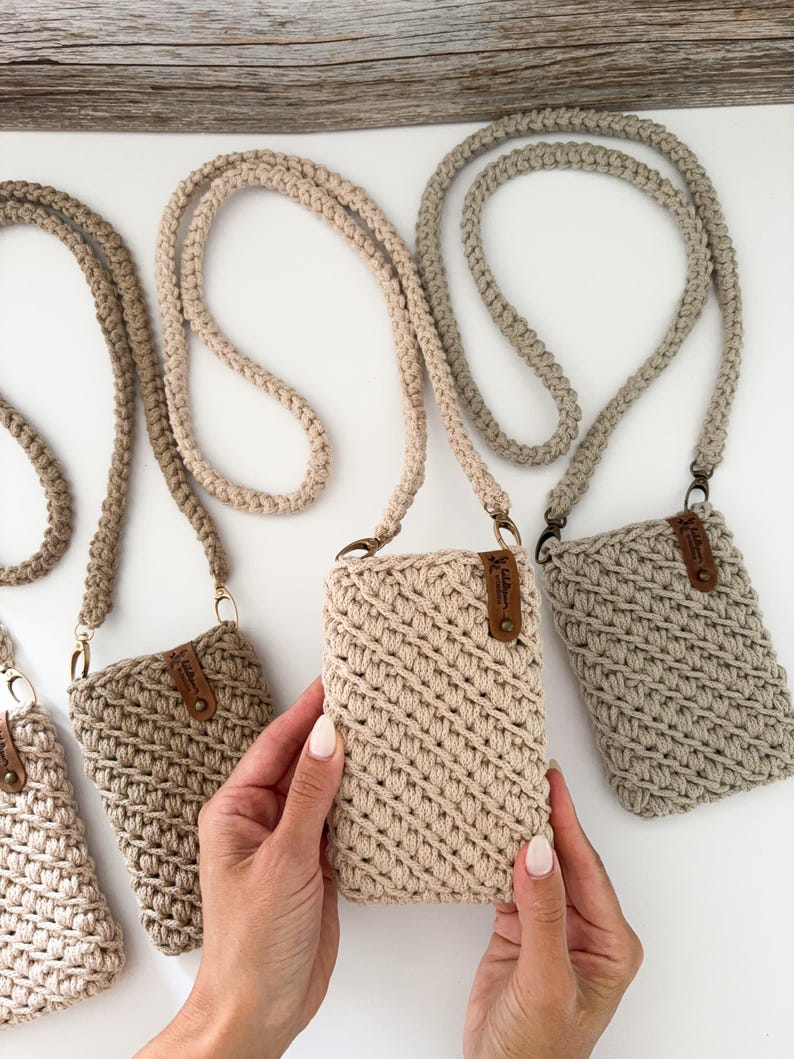 Pu&ograve; includere: Custodie per telefono all'uncinetto in vari colori neutri, tra cui beige e grigio, con cinturini intrecciati coordinati. Ogni custodia presenta una piccola etichetta in pelle e hardware color oro. Esposte su una superficie bianca.