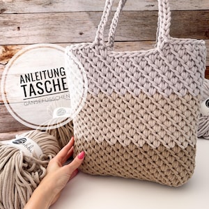 Może przedstawiać: Dwukolorowa szydełkowa torba typu tote w kolorach beżu i jasnoszarości, z teksturowym ściegiem i plecionymi uchwytami. Na okrągłej etykiecie widnieje napis "ANLEITUNG TASCHE". W tle motki przędzy.