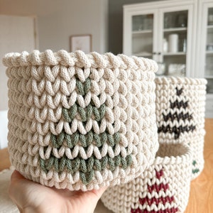 Könnte beinhalten: Drei gehäkelte Körbe mit verschiedenen Weihnachtsbaum-Designs in unterschiedlichen Farben. Die Körbe sind aus naturfarbenem Seil gefertigt und haben ein grünes, braunes und rotes Baumdesign.