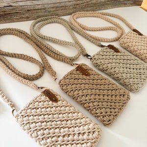 Pu&ograve; includere: Diverse borse a tracolla per telefono all'uncinetto fatte a mano in varie tonalit&agrave; di beige e marrone chiaro. Ogni borsa ha una tracolla intrecciata abbinata e una piccola etichetta in pelle. Le borse sono esposte su una superficie bianca.
