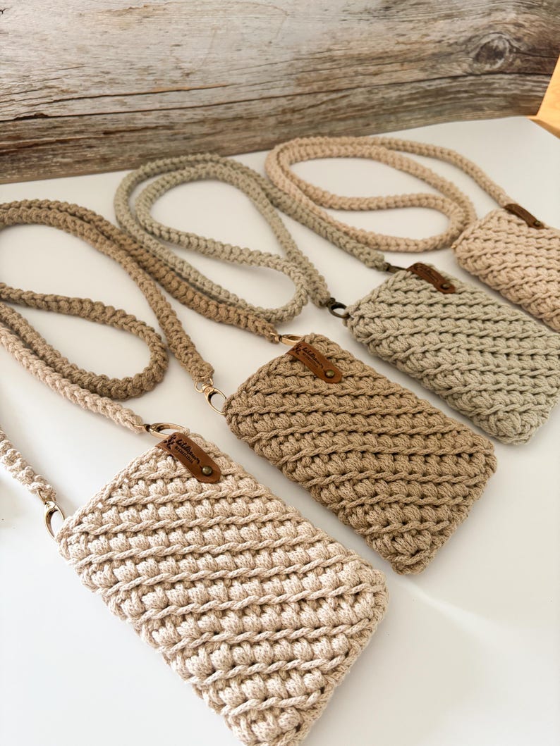 Pu&ograve; includere: Diverse borse a tracolla per telefono all'uncinetto fatte a mano in varie tonalit&agrave; di beige e marrone chiaro. Ogni borsa ha una tracolla intrecciata abbinata e una piccola etichetta in pelle. Le borse sono esposte su una superficie bianca.