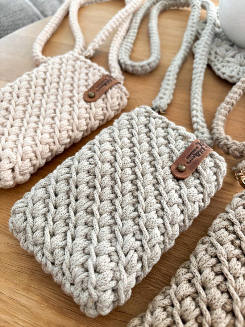 Peut inclure: Gros plan sur plusieurs &eacute;tuis de t&eacute;l&eacute;phone crochet&eacute;s &agrave; la main dans des tons neutres. Chaque &eacute;tui a un motif de point textur&eacute;, une sangle assortie et une petite &eacute;tiquette en cuir. Les &eacute;tuis sont dans des tons beige et gris.