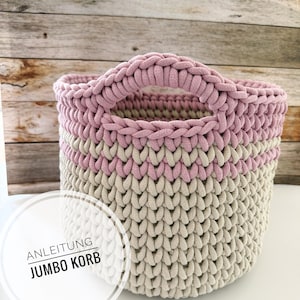 May include: A round, hand-knitted basket with a sturdy handle. The basket features a color-blocked design with a light pink top and a band of light pink in the middle, with a neutral beige color in between. The text "ANLEITUNG JUMBO KORB" is in a white circle.