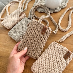 Peut inclure: Plusieurs &eacute;tuis de t&eacute;l&eacute;phone crochet&eacute;s &agrave; la main, dans diff&eacute;rentes nuances de beige et de marron clair, sont pr&eacute;sent&eacute;s sur une surface en bois. Chaque &eacute;tui pr&eacute;sente un motif de point textur&eacute; et une longue sangle tress&eacute;e.