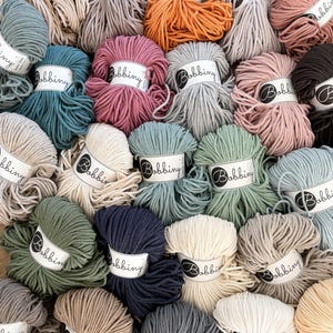 Könnte beinhalten: Eine Sammlung bunter Baumwollseile, jede Spule mit dem Label "Bobbiny" versehen. Das Seil ist kreisförmig angeordnet, wobei die Etiketten nach außen zeigen. Die Farben umfassen Beige, Orange, Rosa, Türkis, Grün, Marineblau und Grau.
