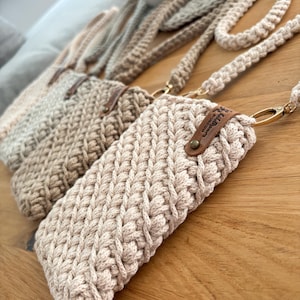 Peut inclure: Gros plan sur trois &eacute;tuis de t&eacute;l&eacute;phone en crochet beige avec de longues lani&egrave;res tress&eacute;es. Les &eacute;tuis ont un aspect textur&eacute; et tiss&eacute; et sont orn&eacute;s de petites &eacute;tiquettes en cuir marron. Les &eacute;tuis sont pos&eacute;s sur une surface en bois.