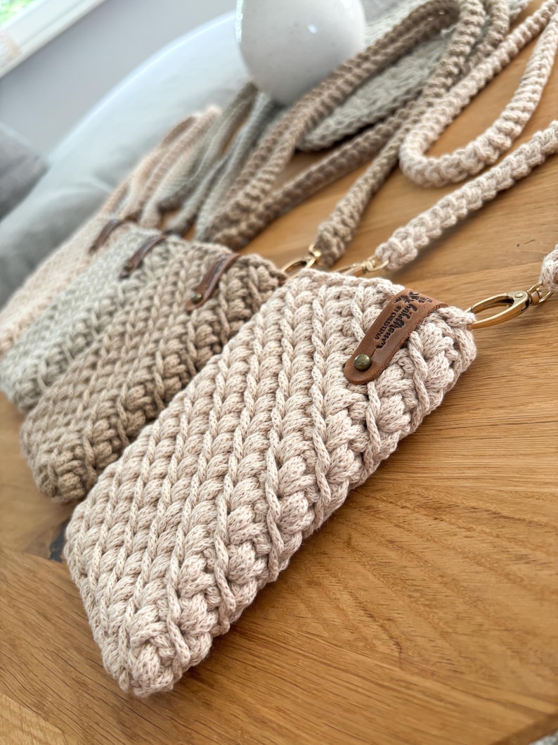 Peut inclure: Gros plan sur trois &eacute;tuis de t&eacute;l&eacute;phone en crochet beige avec de longues lani&egrave;res tress&eacute;es. Les &eacute;tuis ont un aspect textur&eacute; et tiss&eacute; et sont orn&eacute;s de petites &eacute;tiquettes en cuir marron. Les &eacute;tuis sont pos&eacute;s sur une surface en bois.