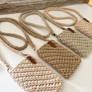 Peut inclure: Plusieurs sacs &agrave; bandouli&egrave;re pour t&eacute;l&eacute;phone en crochet faits &agrave; la main, dans diff&eacute;rentes nuances de beige et de marron clair. Chaque sac est dot&eacute; d'une sangle tiss&eacute;e assortie et d'une petite &eacute;tiquette en cuir. Les sacs sont expos&eacute;s sur une surface blanche.