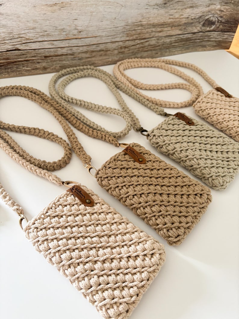 Peut inclure: Plusieurs sacs &agrave; bandouli&egrave;re pour t&eacute;l&eacute;phone en crochet faits &agrave; la main, dans diff&eacute;rentes nuances de beige et de marron clair. Chaque sac est dot&eacute; d'une sangle tiss&eacute;e assortie et d'une petite &eacute;tiquette en cuir. Les sacs sont expos&eacute;s sur une surface blanche.