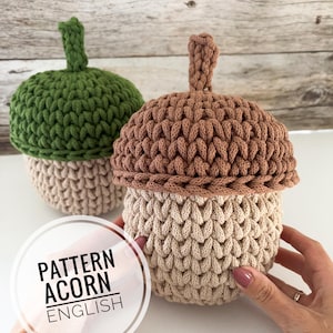 Puede incluir: Dos recipientes de ganchillo con forma de bellota con tapas y tallos texturizados. Una bellota tiene una tapa verde y base beige, mientras que la otra tiene una tapa marrón y base beige. Un gráfico circular blanco muestra el texto PATTERN ACORN ENGLISH.