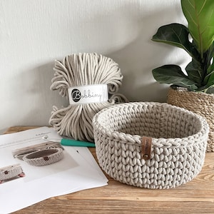 Op de afbeelding: Een beige gehaakte mand met een bruin leren label waarop "HANDMADE" staat. De mand staat op een houten tafel met een bol beige touw en een haaknaald.