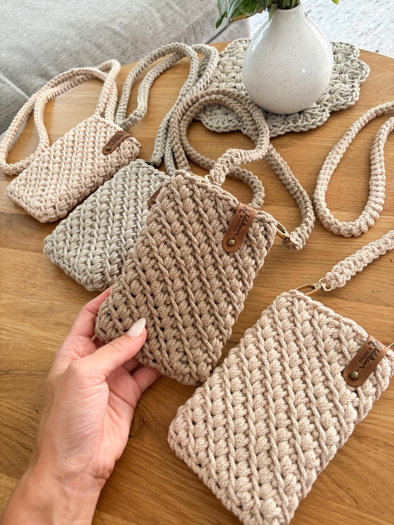 Pu&ograve; includere: Diverse custodie per telefono all'uncinetto fatte a mano in varie tonalit&agrave; di beige e marrone sono esposte su una superficie di legno. Ogni custodia ha un cinturino e una piccola etichetta in pelle. Le custodie presentano diversi motivi a punto all'uncinetto.