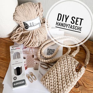 Könnte beinhalten: Ein DIY-Set für eine Makramee-Handytasche. Das Set enthält einen Knäuel beiges Bobbing-Garn, einen Häkelhaken, eine fertige Handytasche und weiteres Zubehör. Der Text "DIY SET HANDYTASCHE" ist sichtbar.