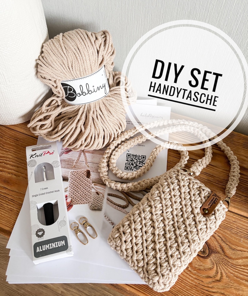 Pu&ograve; includere: Un kit fai-da-te per borsa per telefono in macram&egrave;. Il kit include un gomitolo di filato Bobbing beige, un uncinetto, una borsa per telefono finita e altri accessori. Il testo "DIY SET HANDYTASCHE" &egrave; visibile.