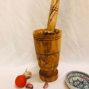 Op de afbeelding: Een houten vijzel en stamper set van olijfhout. De vijzel is cilindrisch met een uitlopende basis en de stamper heeft een afgeronde handgreep. Een rode tomaat, knoflookteentjes en een kleine decoratieve kom zijn ook aanwezig.