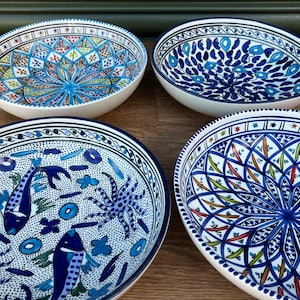 May include: Four white ceramic bowls with detailed blue patterns. The bowls display diverse designs, including floral and geometric motifs. The rims are outlined in blue, and the bowls are on a wooden surface.