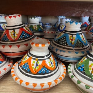 May include: A selection of hand-painted ceramic tagine pots. Each pot has a conical lid and a round base, decorated with patterns in red, blue, orange, and green. The pots are displayed on a wooden shelf, with each pot's measurements in cm.
