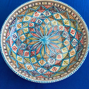 May include: A round, hand-painted ceramic plate with a vibrant, geometric design. The plate features a central starburst pattern in shades of blue, red, yellow, and green, surrounded by intricate border details. The plate is approximately 30 cm in diameter.