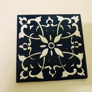 May include: Square, navy blue tile with a white floral pattern. The design includes a central flower, radiating petals, and curved lines to corner details. The tile is probably ceramic or porcelain.