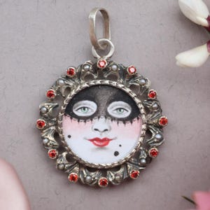May include: A silver pendant with a painted face design. The face has a black mask, red lips, and green eyes. The pendant is surrounded by red gemstones and floral accents. A unique piece of jewelry.