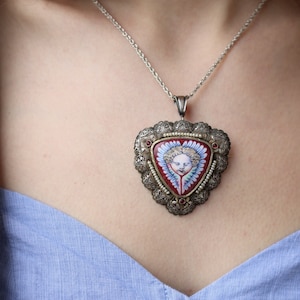 May include: A silver chain necklace with a triangular pendant featuring a red enamel background and a painted image of an angel with blue wings. The pendant is decorated with silver filigree and pearl accents.