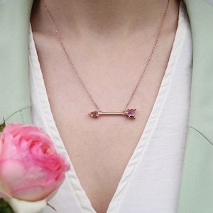 Może przedstawiać: Złoty naszyjnik z wisiorem w kształcie strzały z różowym kamieniem szlachetnym. Naszyjnik nosi osoba w białej koszuli i jasnozielonym blazerze.