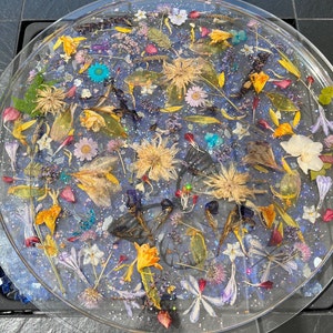 May include: A round, clear resin tray with a variety of pressed flowers and leaves embedded in it. The flowers are in shades of pink, purple, yellow, and white, and the leaves are green and brown. The tray is filled with glitter, giving it a sparkly appearance.