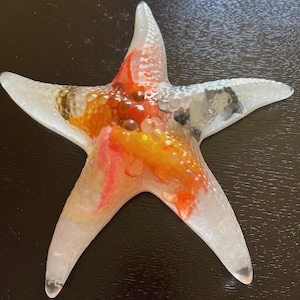 May include: A white glass starfish with orange, red, and yellow accents. The starfish has a textured surface with small bubbles.
