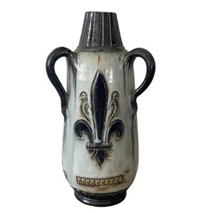 Peut inclure: Un vase en céramique blanche avec un motif de fleur de lys bleu et noir. Le vase a deux anses et une surface texturée.