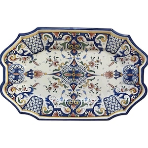 Pode incluir: Uma bandeja de servir retangular de cerâmica com borda ondulada. A bandeja apresenta um fundo branco com um padrão floral e geométrico detalhado em azul, laranja e verde. A borda é azul escuro.
