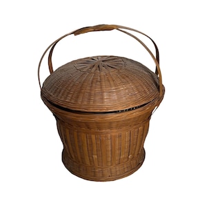 以下が含まれることがあります： ふたと2つの取っ手のある、茶色の籐で編まれたバスケット。バスケットは自然素材で作られており、素朴な外観をしています。