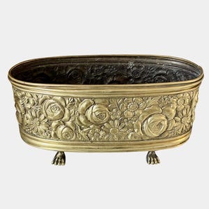 Puede incluir: Una jardinera ovalada de latón con un patrón floral en relieve de rosas y margaritas en su superficie exterior. El recipiente se apoya sobre cuatro pequeñas patas en forma de garra y tiene un acabado interior oscuro y envejecido.