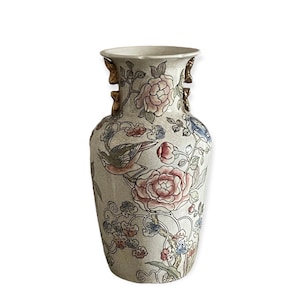 Puede incluir: Un jarrón de cerámica blanca con un diseño floral y de pájaros. El jarrón tiene detalles dorados y una abertura ligeramente acampanada.