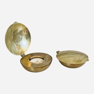 Puede incluir: Un candelabro esférico de color dorado con tapa abisagrada. El soporte está abierto, revelando una vela blanca encendida en el interior. La tapa está separada y colocada a un lado. El candelabro tiene un diseño simple y elegante.