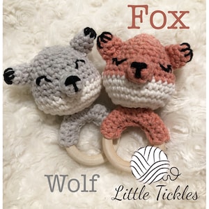 May include: A gray crocheted wolf and a pink crocheted fox attached to a wooden teething ring. The ring is decorated with crocheted details in gray and pink. The text "Wolf" and "Fox" are written below the animals. The text "Little Tickles" is written below the ring.