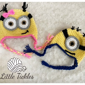 May include: Two crocheted Minion hats made with yellow, blue, black, white and pink yarn. The hats have a braided cord and a single eye on each hat. The hats are on a white fluffy surface. The text "Little Tickles" is visible in the bottom centre of the image.