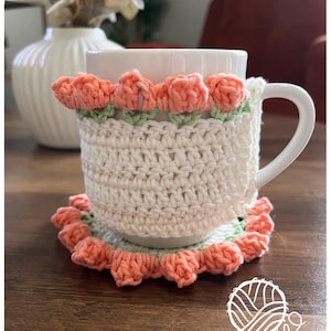 May include: A white ceramic mug with a crocheted cozy and coaster. The cozy and coaster are cream-colored with peach-colored flower accents and green leaves. The mug and accessories are on a wooden surface. The logo "Little Tickles" is visible.