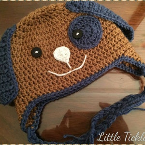 May include: A brown and blue crocheted hat shaped like a dog with black button eyes and a white crocheted nose. The hat has a blue crocheted brim and a long blue crocheted tie.