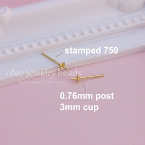 May include: Two gold earring posts, each with a 3mm cup and a 0.76mm post. The posts are stamped with "750". The background is a light pink surface with a white decorative border. The text "dee jewelry beads" is visible.