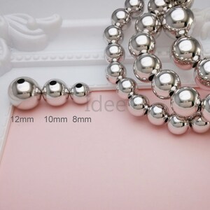 May include: Three silver metal beads of different sizes, 12mm, 10mm, and 8mm, with a strand of larger silver beads in the background.