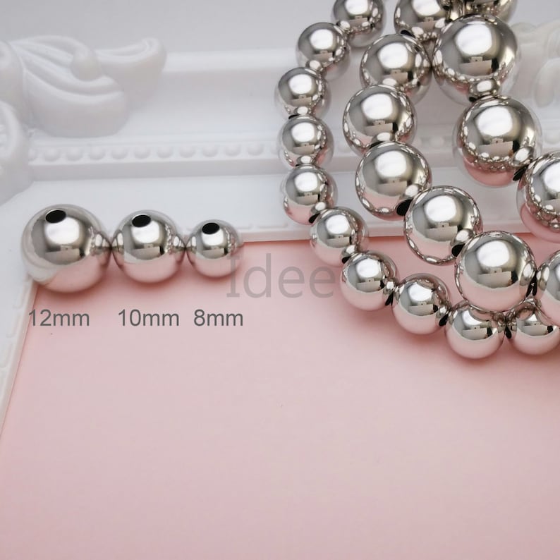 May include: Three silver metal beads of different sizes, 12mm, 10mm, and 8mm, with a strand of larger silver beads in the background.