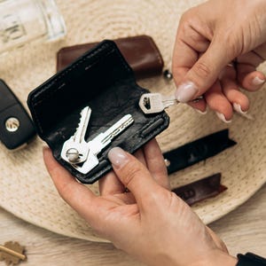May include: Black leather key holder with silver keys inside. A person is holding the key holder, with other keys, a car key fob, and a bottle of cologne nearby. The key holder is open, revealing the keys.