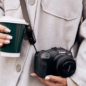 May include: A black Canon EOS camera with a lens is held. The camera is attached to a brown leather strap. A dark green coffee cup with a white lid is held in the other hand. The person is wearing a light grey jacket.