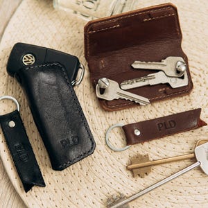 May include: An assortment of leather keychains and key holders in black and brown. The keychains and key holders have the letters "PLD" embossed on them. A car key is also present.