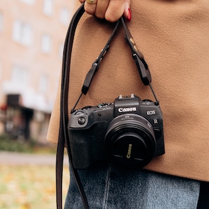 Może przedstawiać: Czarny aparat Canon EOS R z osłoną obiektywu, zawieszony na brązowym skórzanym pasku. Aparat jest trzymany na tle beżowego płaszcza i niebieskich dżinsów. Widoczne jest logo Canon.