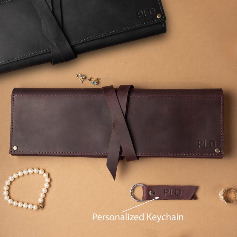 May include: A dark burgundy leather roll-up case with a tie closure and the embossed letters PLD, accompanied by a matching leather keychain with the letters PLD, a pearl bracelet, a gold ring, and a black leather roll-up case with PLD.